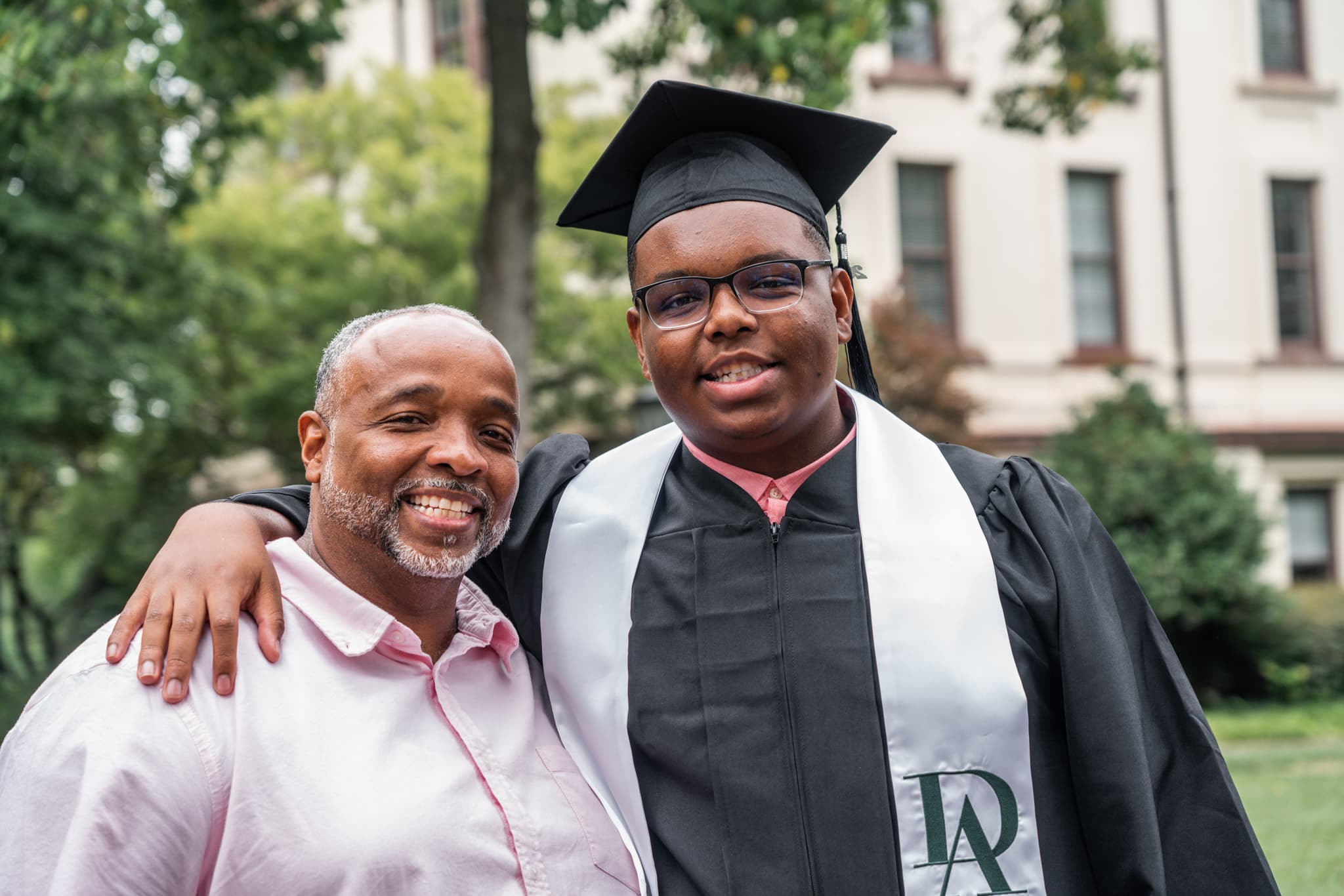 Two individuals are smiling for the camera, one wearing a graduation cap and gown, and the other dressed in a pink shirt. They appear to be celebrating a graduation event