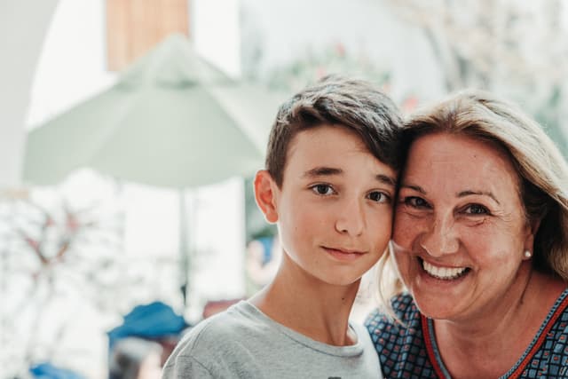 A smiling older woman embraces a young boy, both looking at the camera, with a blurred indoor background suggesting a home environment