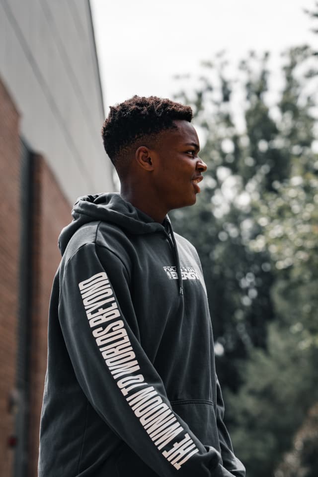 A young man in a hooded sweatshirt stands outdoors, looking to the side with a thoughtful expression. The hoodie features prominent lettering along the sleeve