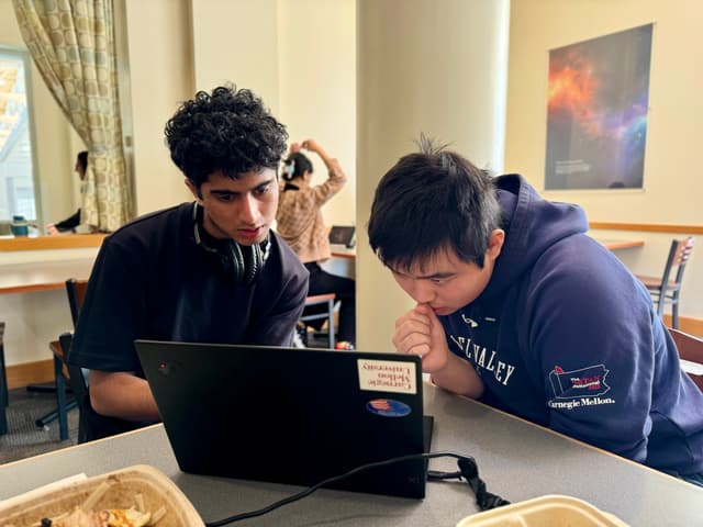 Two individuals are focused on a laptop screen, with one person pointing at the screen, possibly discussing something. They are seated at a table with food containers, suggesting a casual working or study environment