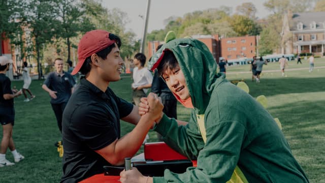 Two individuals are engaged in an arm wrestling match on a table outdoors, with onlookers in the background