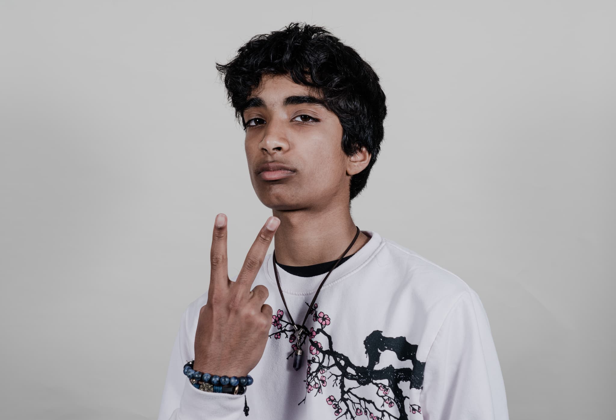 A young man with curly hair is posing against a grey background, wearing a white graphic t-shirt and making a peace sign with his right hand