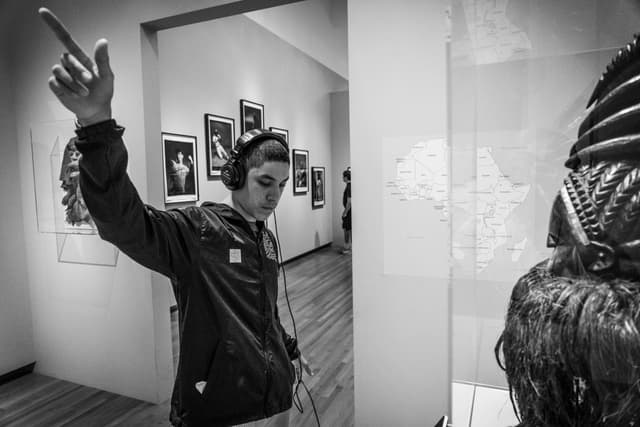 A person wearing headphones is gesturing with one arm raised in an art gallery, with photographs on the wall and a sculpture visible in the foreground. The image is in black and white