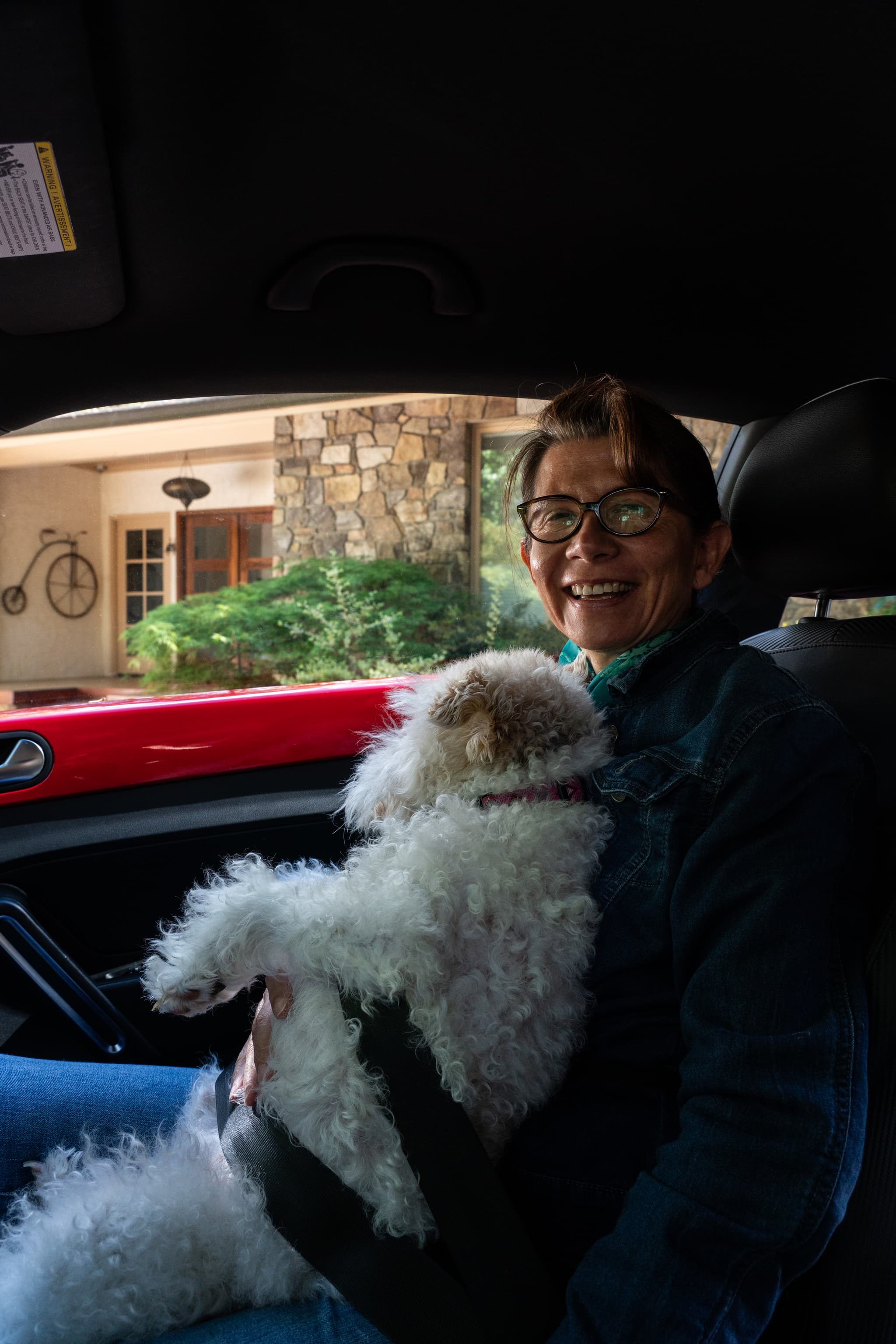 A smiling person wearing glasses is seated in a car, holding a white fluffy dog on their lap