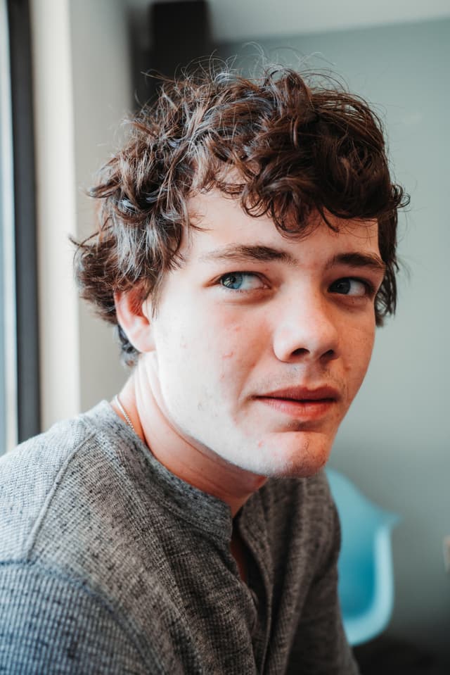 A young man with curly hair looks at the camera, wearing a grey shirt, with a blurred background