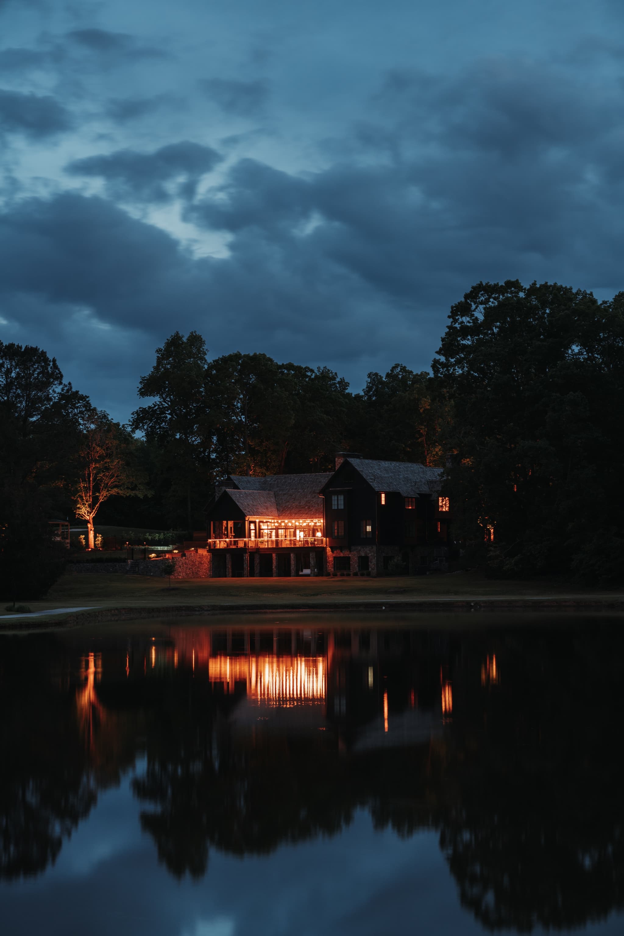 A house with illuminated windows is reflected on a body of water under a twilight sky