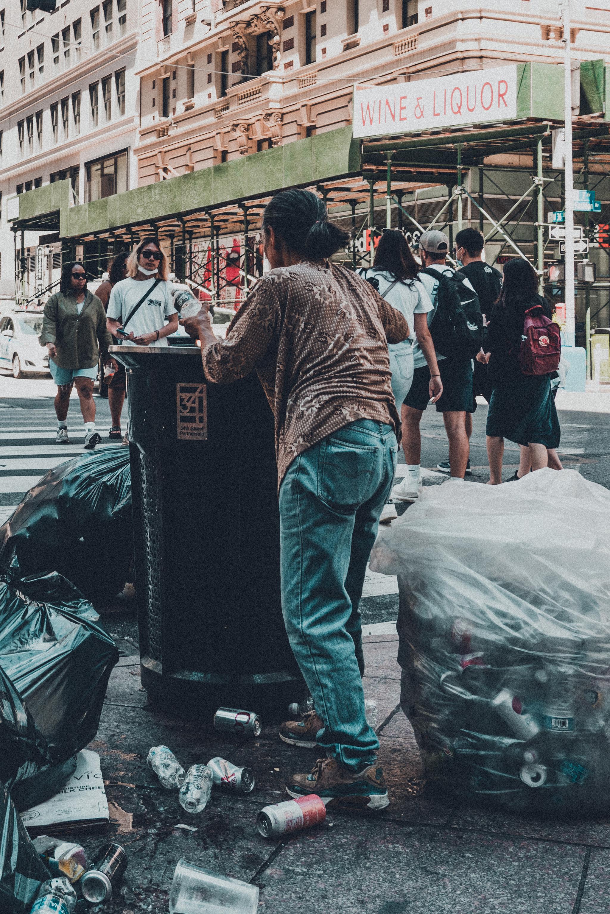 A person stands on a skateboard while rummaging through a trash bin on a city street, with bags of garbage nearby and pedestrians in the background