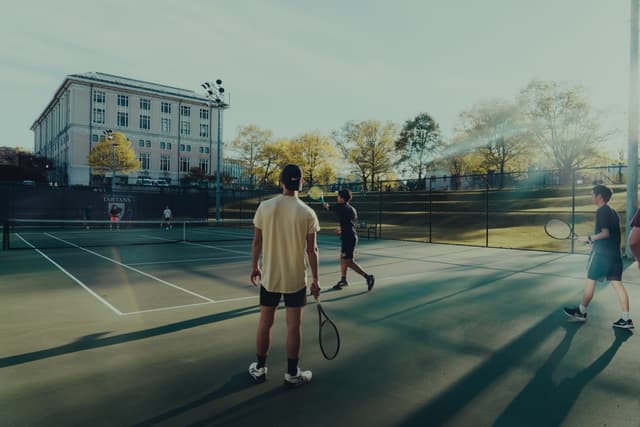 Four individuals are playing doubles tennis on an outdoor court, with a building and trees in the background. The sunlight suggests it's either early morning or late afternoon