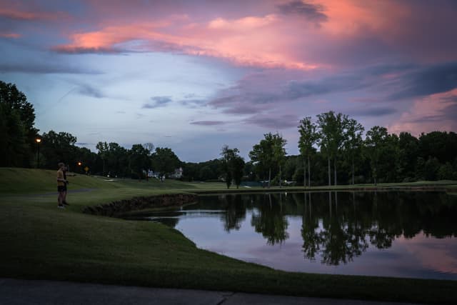 A serene twilight scene over a golf course with a colorful sky reflected in a still pond, and a solitary golfer in the distance