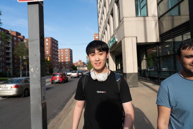 Two individuals are walking on a sidewalk in an urban setting with buildings and parked cars in the background. The person in the foreground is smiling at the camera and wearing a black t-shirt and headphones around their neck, while the other person is partially visible to the right