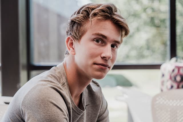 A young man with short hair, looking to the side, seated indoors with a window in the background