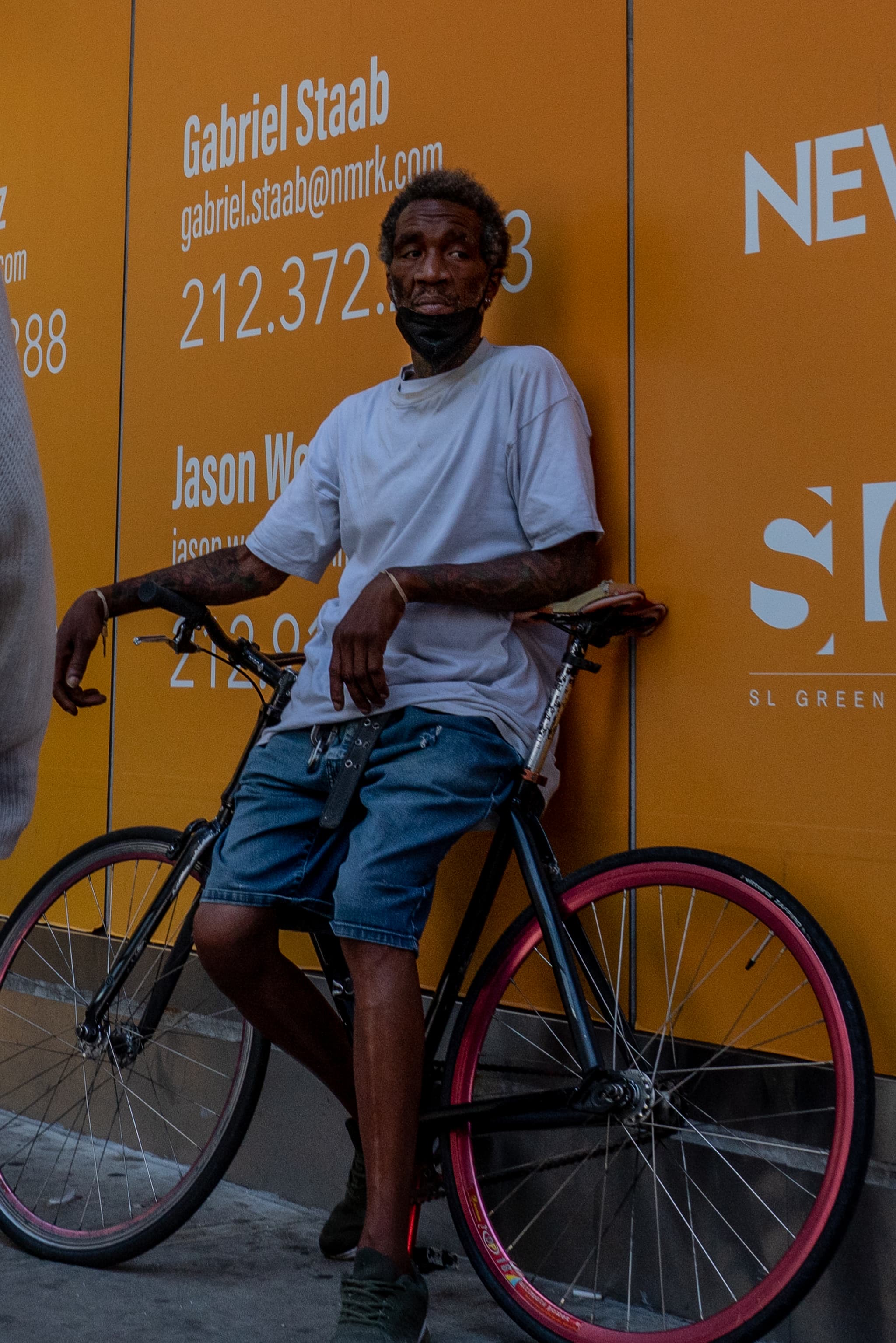 A man leans against a vibrant orange wall with text and phone numbers, holding onto a bicycle with one red wheel and one black wheel. He's wearing a white t-shirt, blue shorts, and sunglasses, and has a relaxed posture