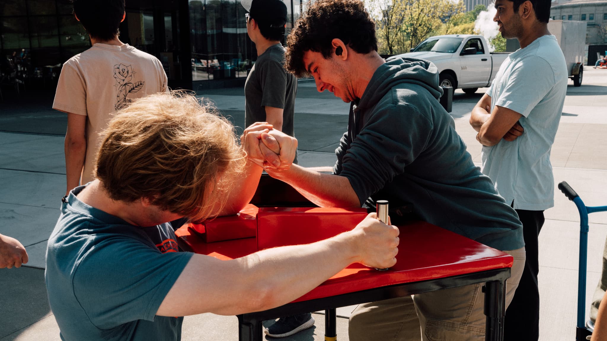 Two men are engaged in an arm wrestling match on a red table outdoors, with onlookers observing the competition