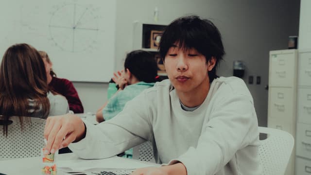 A young man sits at a table, looking directly at the camera with a playful expression and puckered lips, as if blowing a kiss or holding a small object between them. In the background, other individuals are engaged in conversation, with whiteboards and office supplies suggesting a classroom or office setting