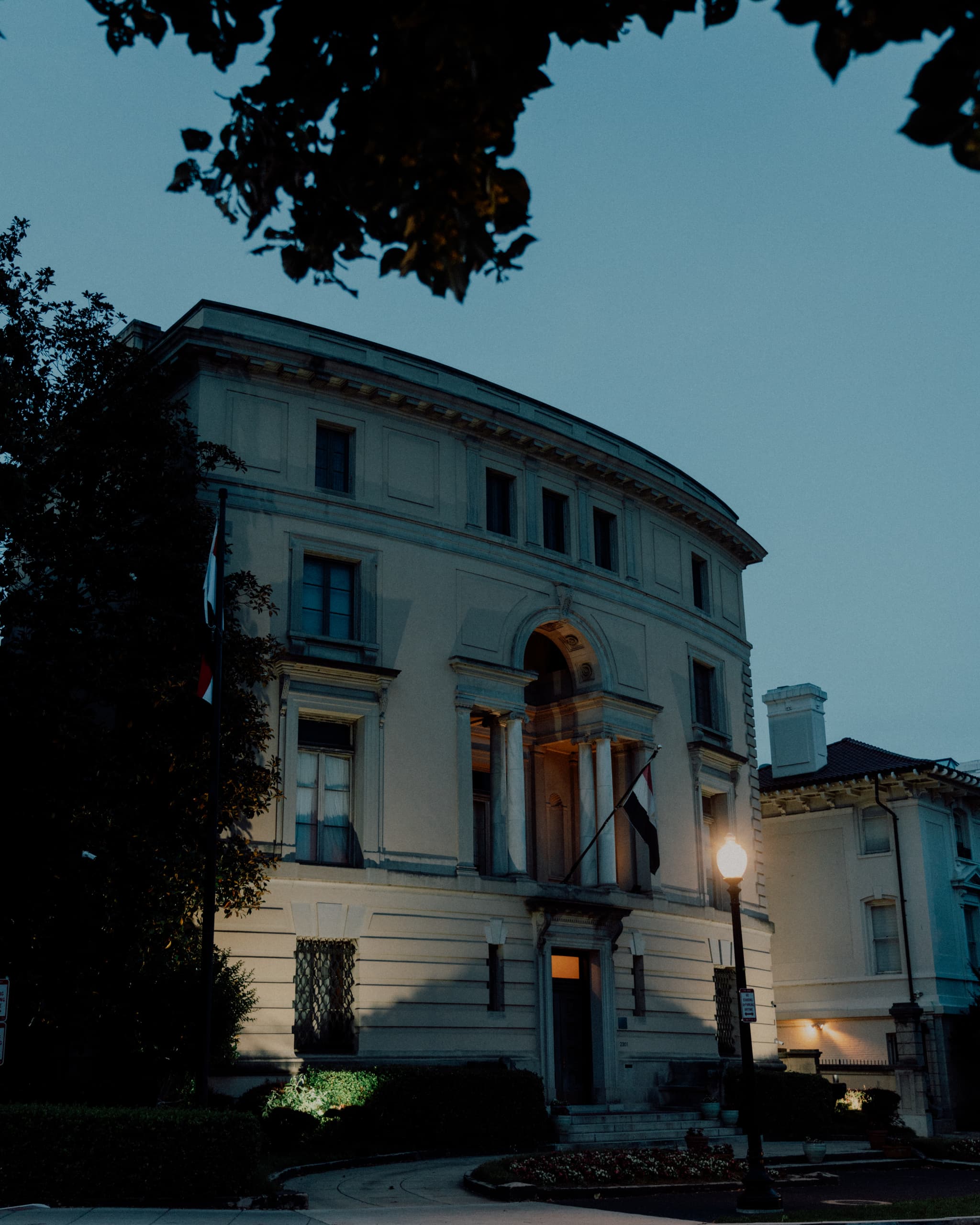 A stately building captured at dusk, with lights illuminating the entrance and windows, framed by silhouetted foliage