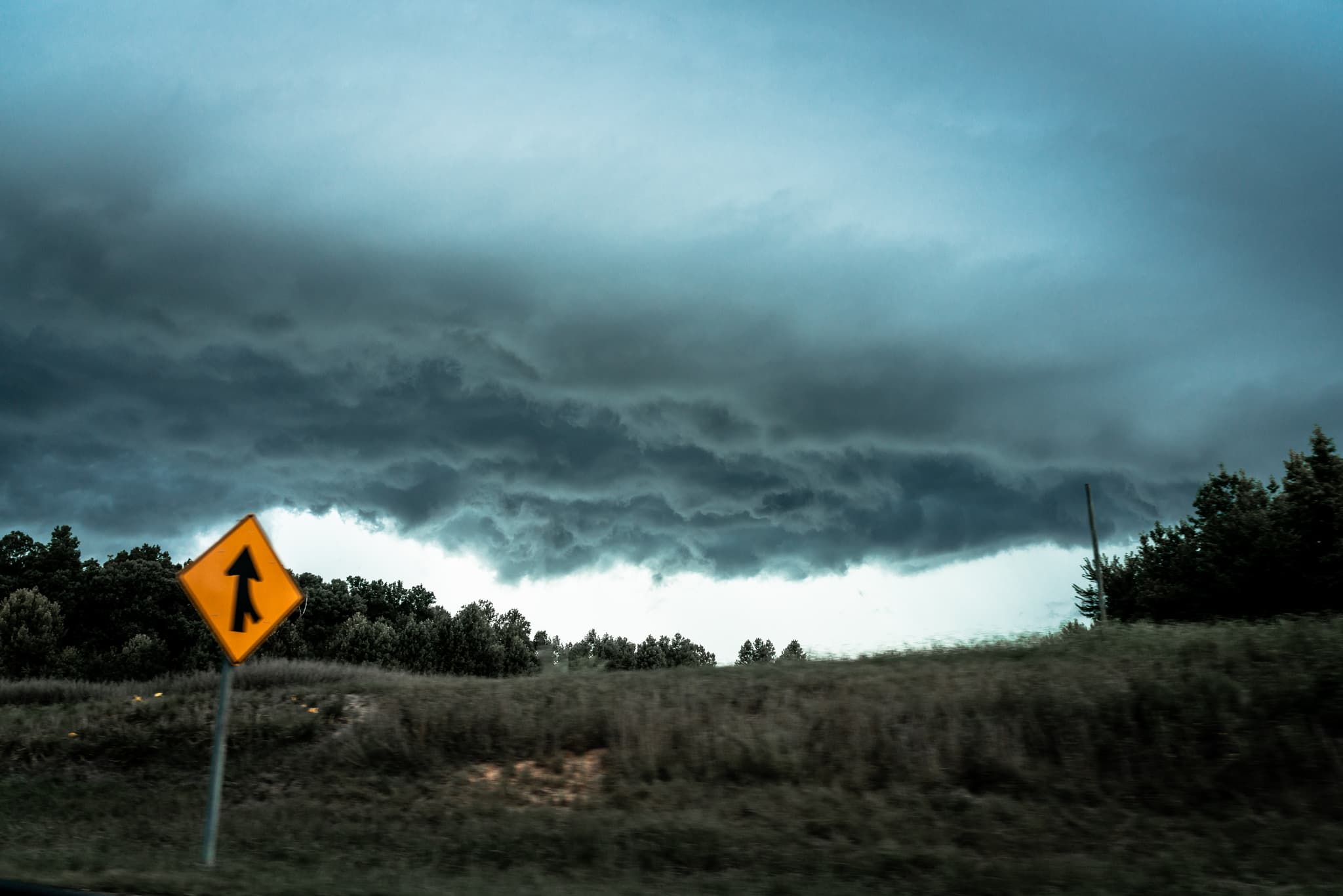 A dark, stormy sky looms over a landscape with a yellow road sign indicating a merge or lane coming together