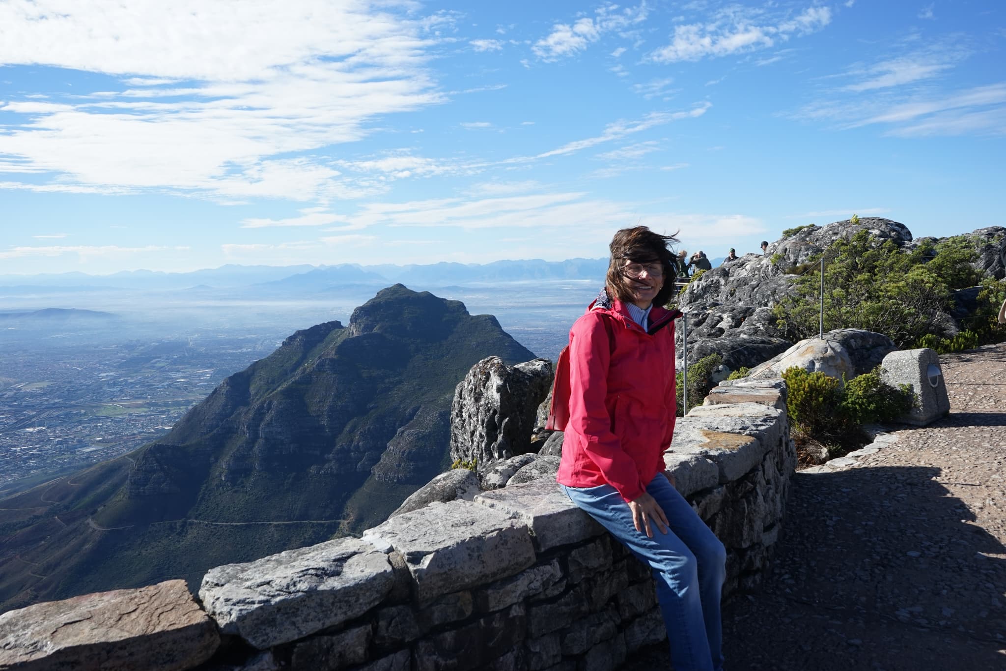 Monica Lopez-Vazquez sits on a stone wall at a mountain summit, with rugged peaks and a vast landscape stretching into the distance