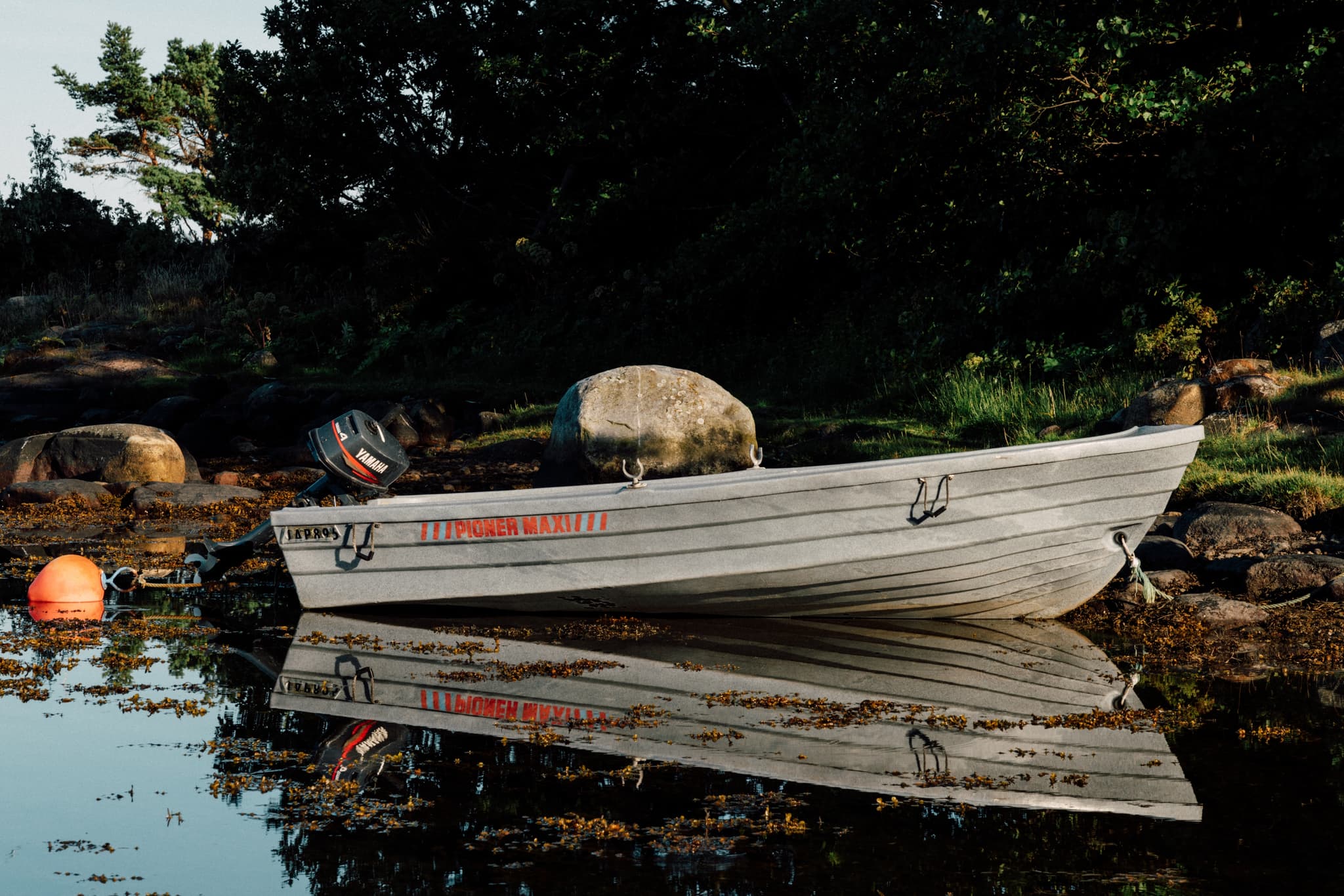 A weathered rowboat rests on a rocky shore beside calm water, with greenery in the background