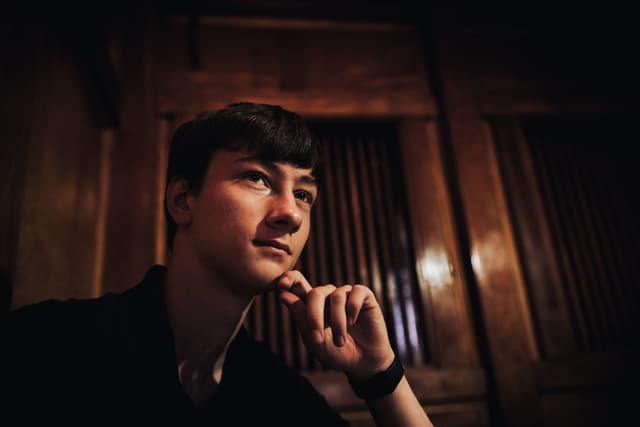 A contemplative young man, Daniel Arcidiacono, with his hand on his chin, set against a dark, wooden background with soft lighting