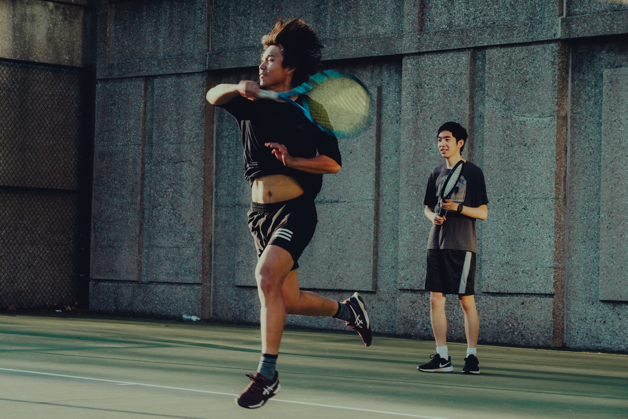 Two people playing tennis outdoors, with one person in mid-air hitting the ball and the other person standing in the background watching