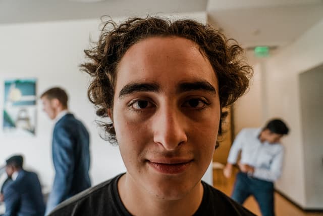 A close-up of a young man with curly hair smiling at the camera, with two other individuals in the background