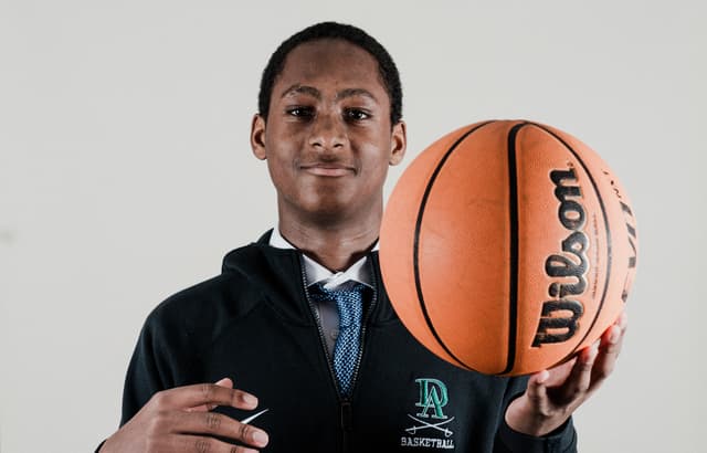 A young man in a dark blazer holds a basketball, looking at the camera with a slight smile