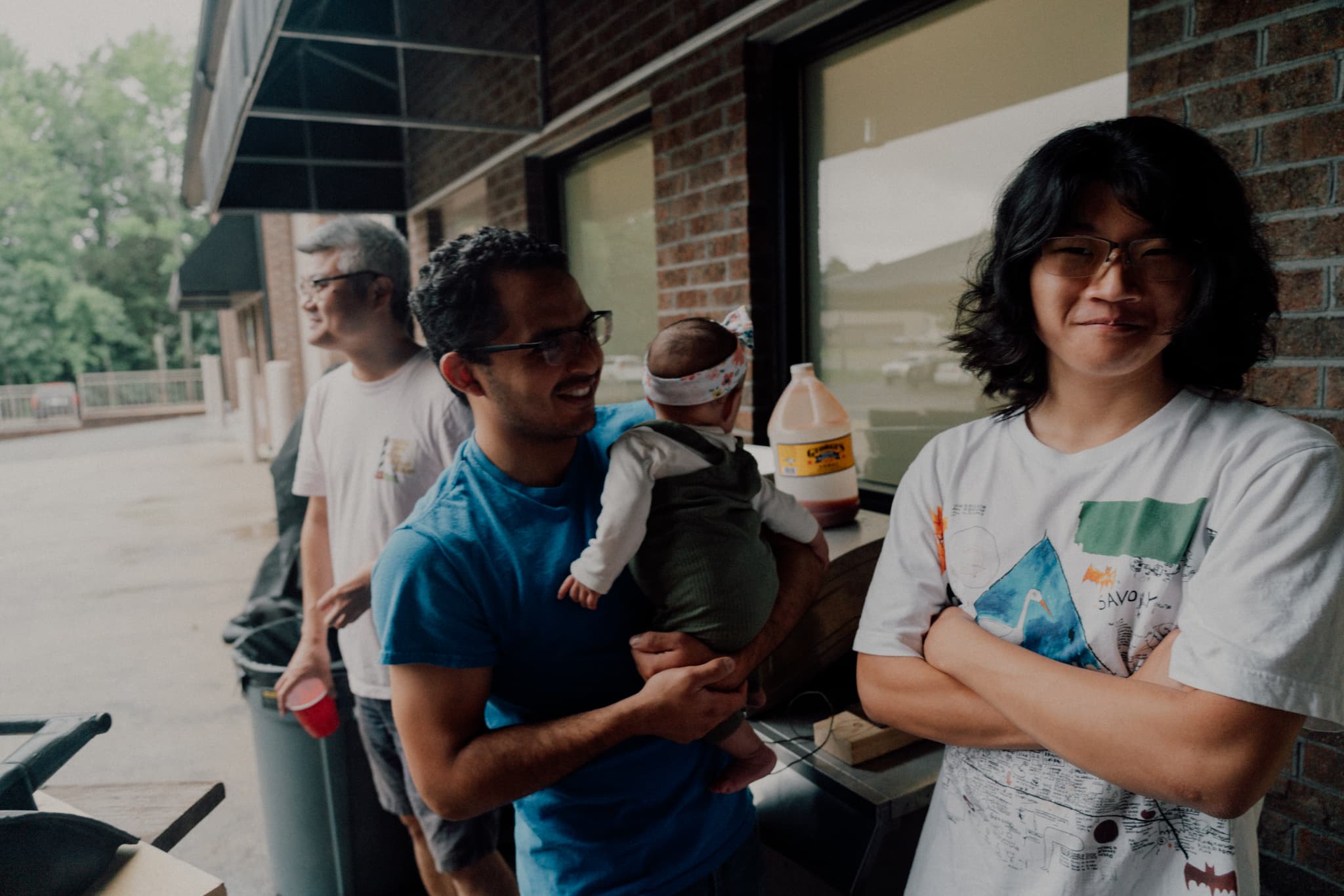 A group of people stands outside a building, with one person holding a child and another smiling at the camera