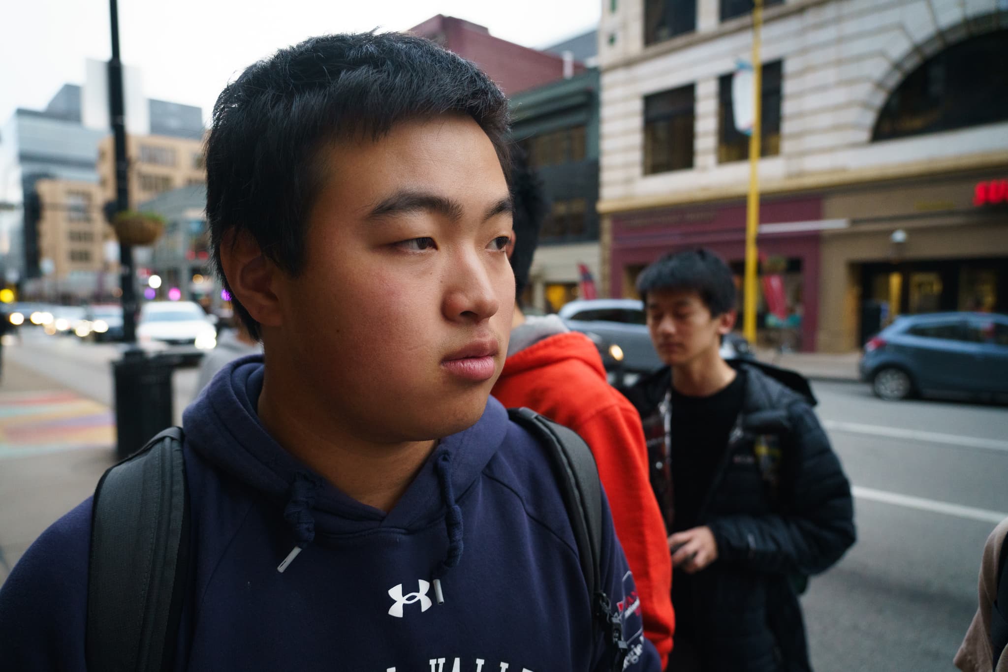 A young person wearing a dark jacket with a backpack, looking to the side on a city street with buildings and another individual in the background