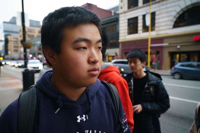 A young person wearing a dark jacket with a backpack, looking to the side on a city street with buildings and another individual in the background