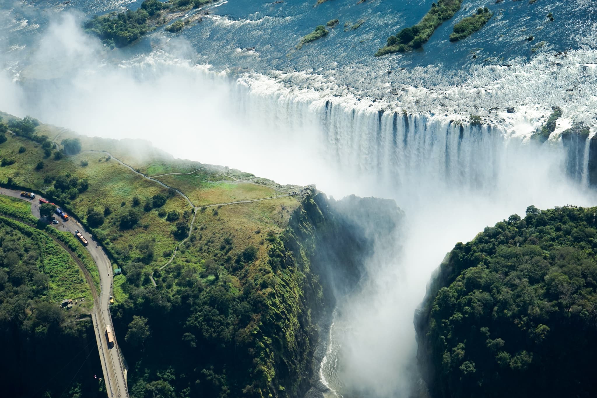 An aerial view of a majestic waterfall with a large volume of water plunging down, generating mist, surrounded by lush greenery and a winding road nearby