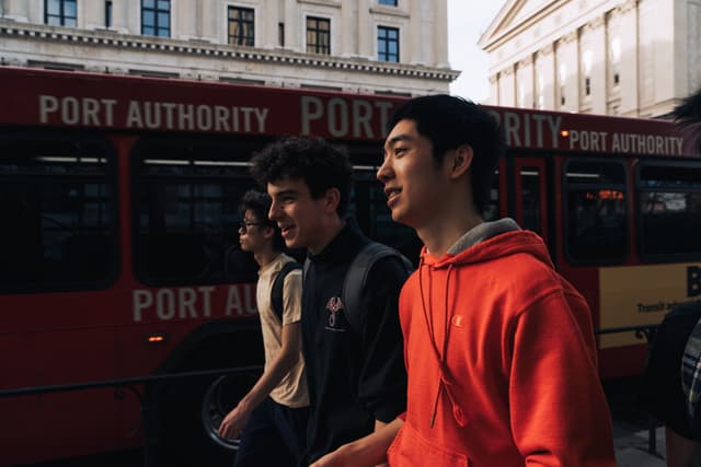 Three individuals are standing in front of a red bus with the words Port Authority visible on its side. The person in the foreground is wearing a red hoodie and looking to the side, with sunlight illuminating half of his face. The background features a building with classical architecture, suggesting an urban setting