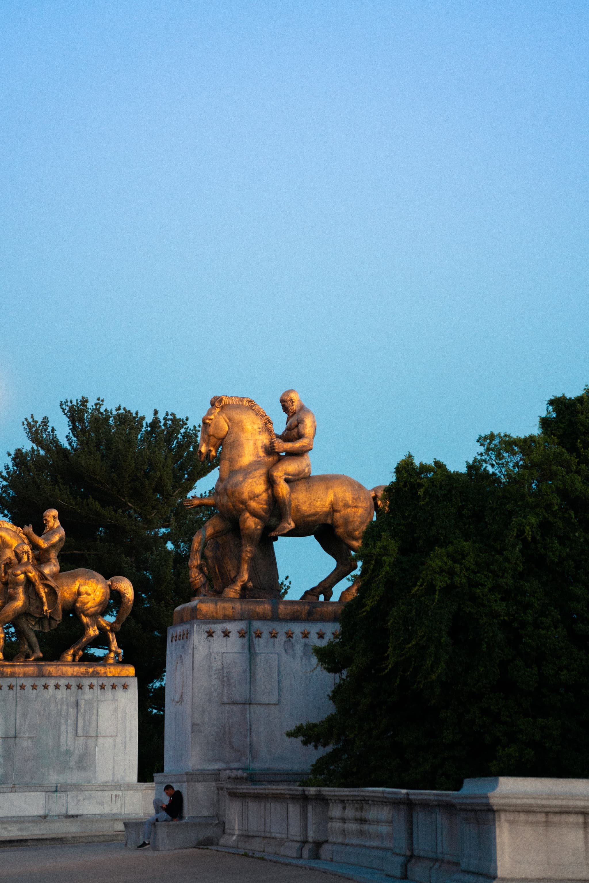 Bronze equestrian statues are perched atop a stone structure in the early evening, with a clear sky in the background and a solitary figure walking below