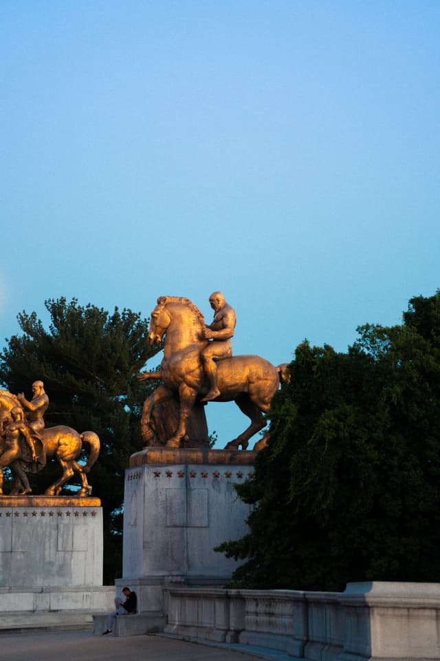 Bronze equestrian statues are perched atop a stone structure in the early evening, with a clear sky in the background and a solitary figure walking below