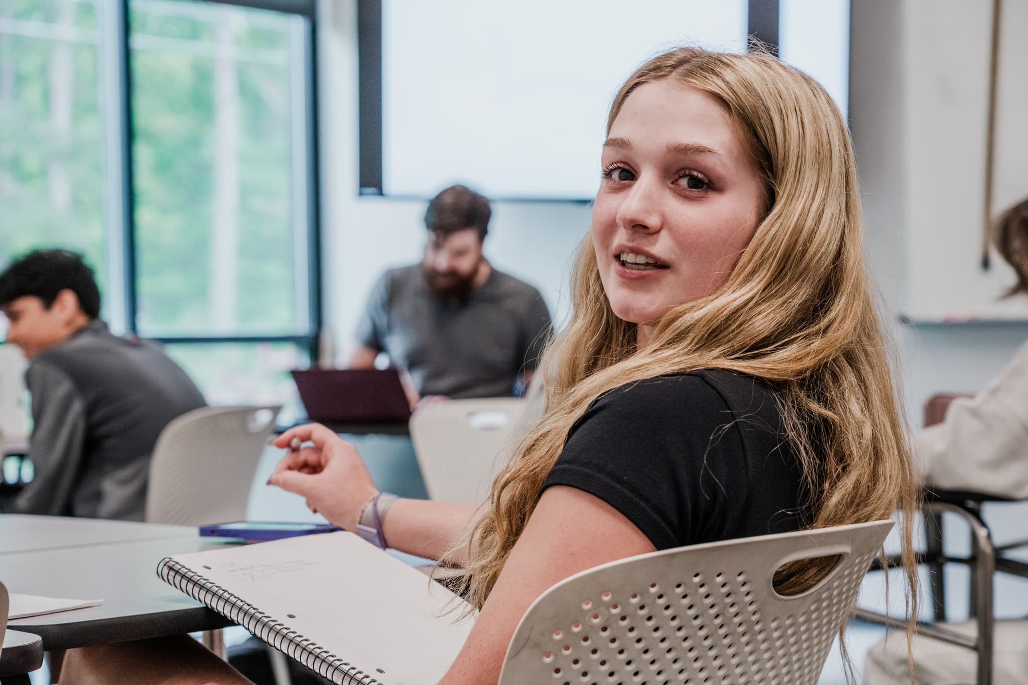 A young woman glances back at the camera with a slight smile, seated in a classroom setting with other students and laptops visible in the background