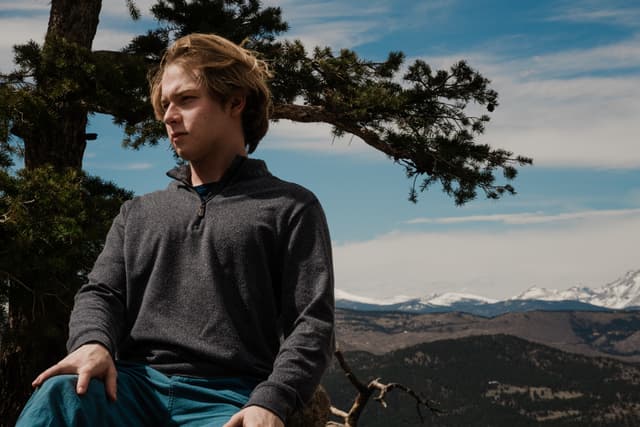 A young man sits outdoors with a backdrop of mountains and a clear sky, partially shaded by a tree