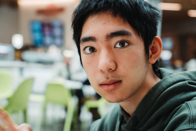 A young man with dark hair looks directly at the camera, a hint of a smile on his face, with a blurred background suggesting an indoor setting with green chairs