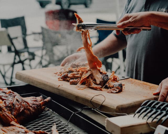 A person carves meat on a wooden board with tongs, with a barbecue grill and outdoor seating in the background