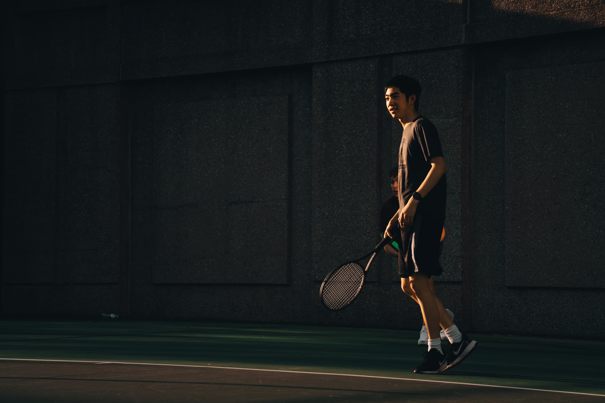 A person walks on a tennis court holding a racket, with the scene cast in shadow and sunlight