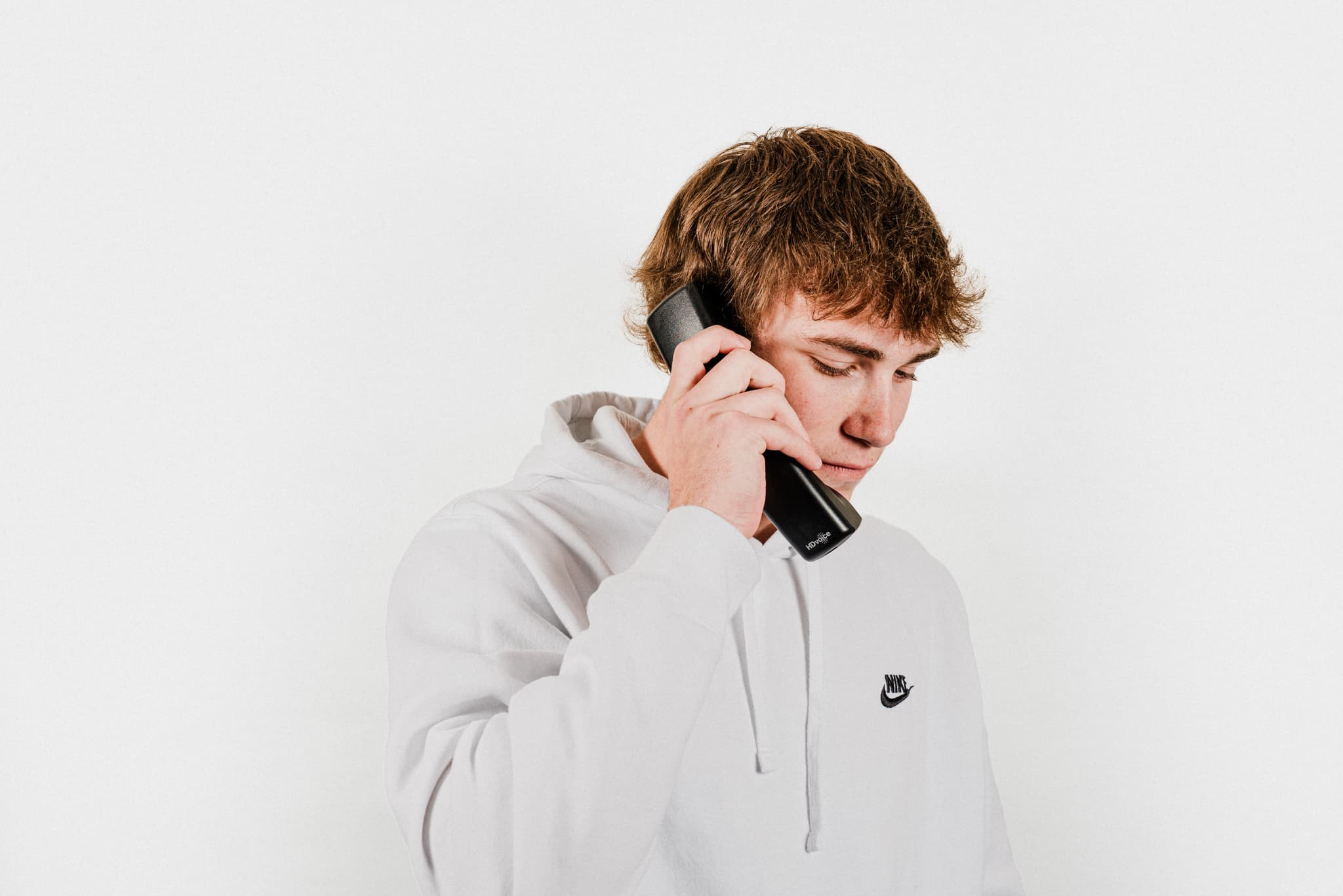 A young man with curly hair, wearing a light-colored hoodie, is holding a mobile phone to his ear, seemingly engaged in a conversation. The background is plain and white