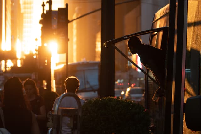 A silhouette of a person stepping off a bus at sunset, with the warm glow of the sun casting long shadows on an urban street