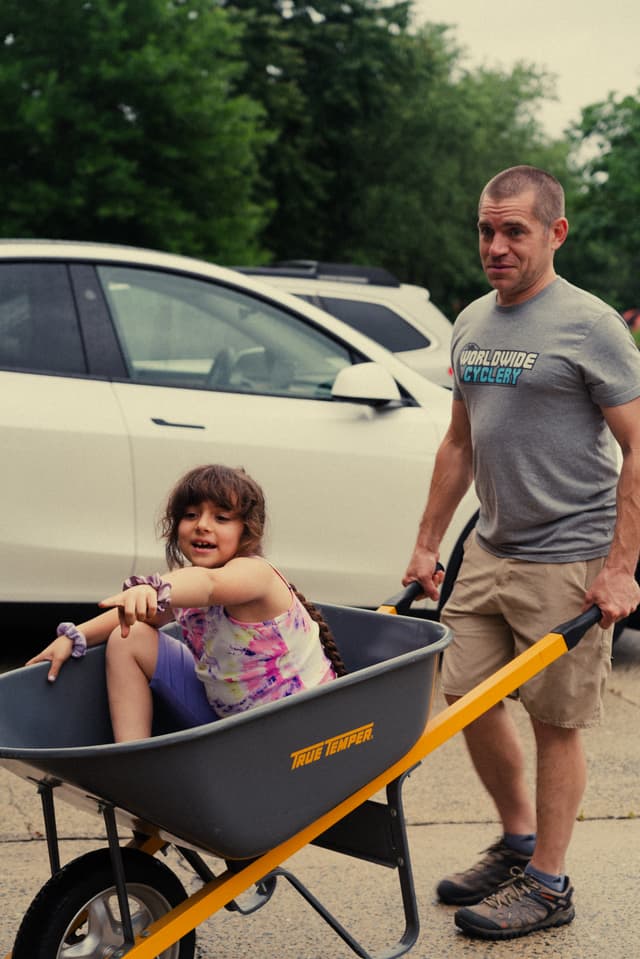 A man pulls a young girl sitting in a wheelbarrow, both appearing to enjoy a leisurely moment outdoors