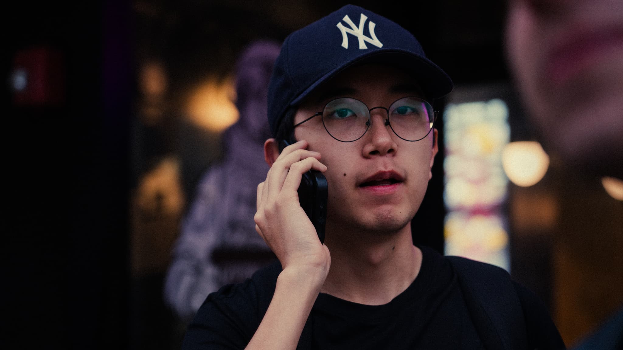 A young man wearing glasses and a New York Yankees cap is talking on a mobile phone, with a blurred background suggesting an urban, possibly nighttime setting