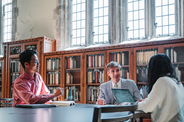 Three individuals (John Rose, Ming Wilson) are seated at a table in a library with large windows and bookshelves in the background, engaged in what appears to be a discussion or study session