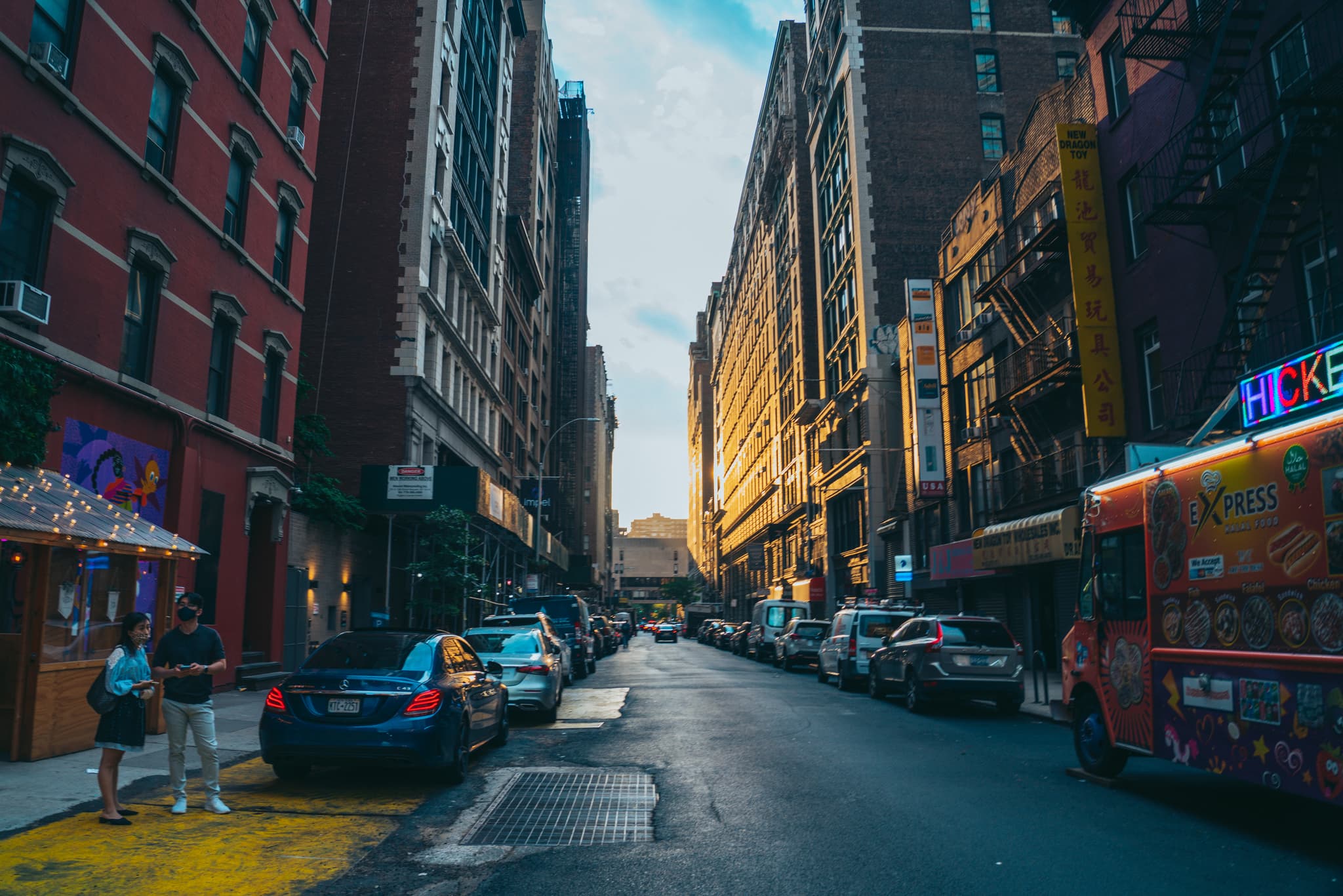 A narrow city street lined with tall buildings, parked cars, and a few pedestrians, bathed in the warm glow of a setting or rising sun