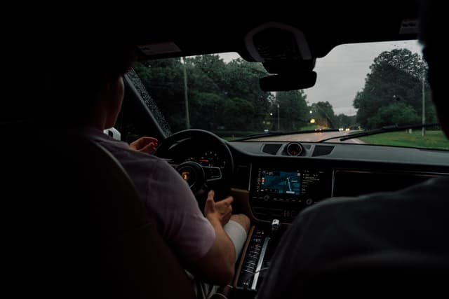 A view from the backseat of a car, capturing the driver and passenger in the front seats with the dashboard and windshield visible, as they drive down a road lined with trees