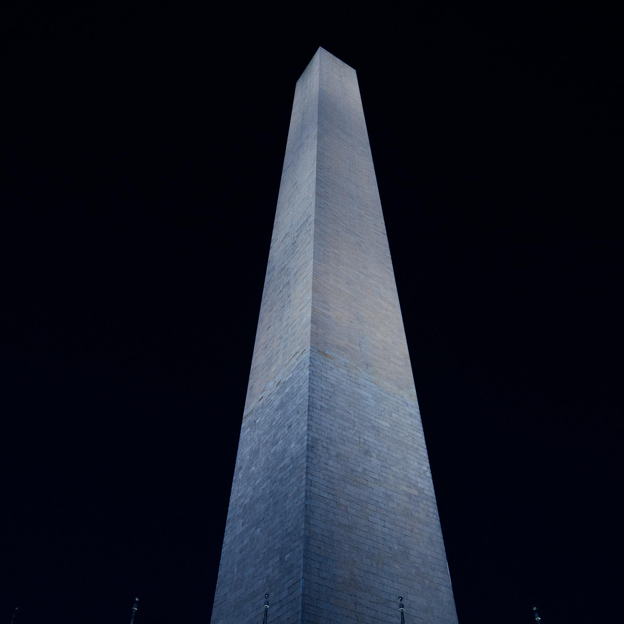 A towering obelisk rises against a dark night sky, illuminated from below to highlight its structure and texture