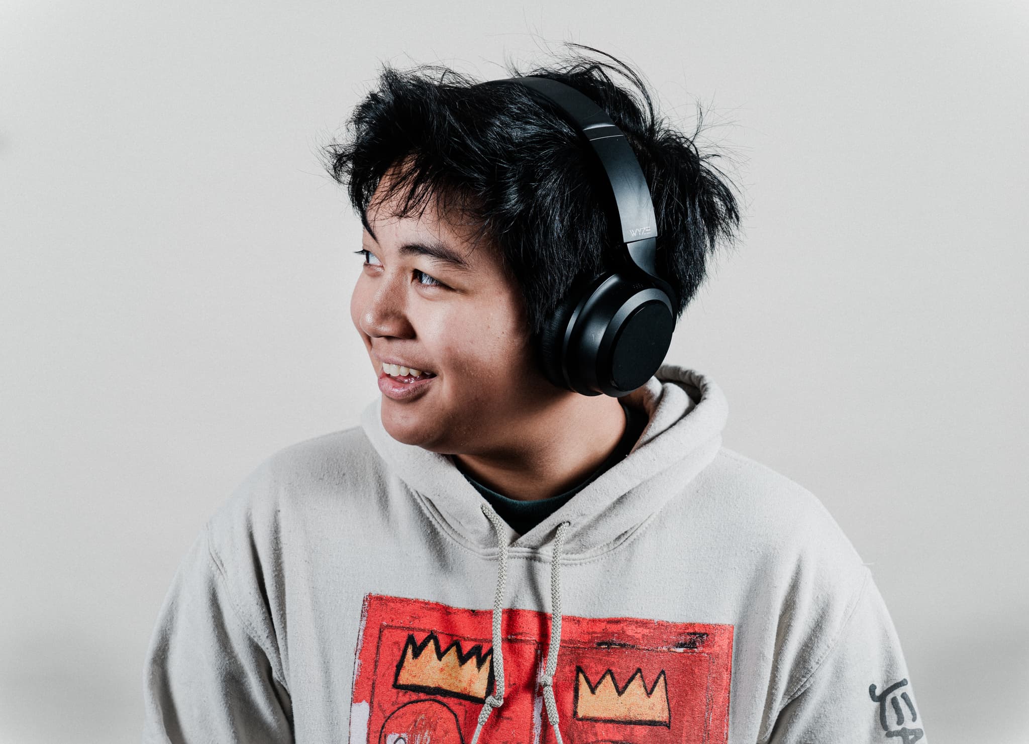 A smiling person wearing a hoodie and headphones around their neck, looking to the side against a light gray background