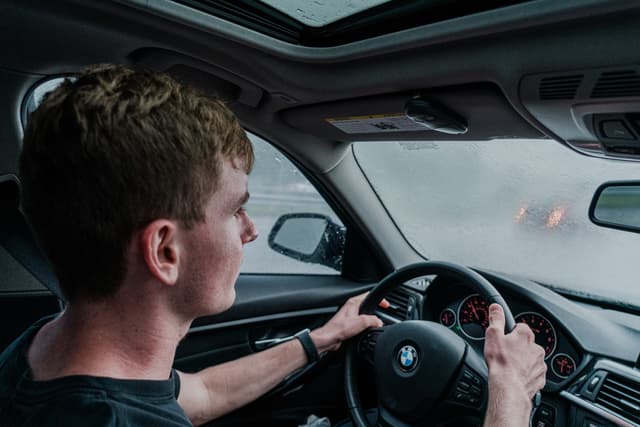 A young man is driving a car, focusing on the road ahead. He's behind the wheel of a vehicle with a BMW logo on the steering wheel