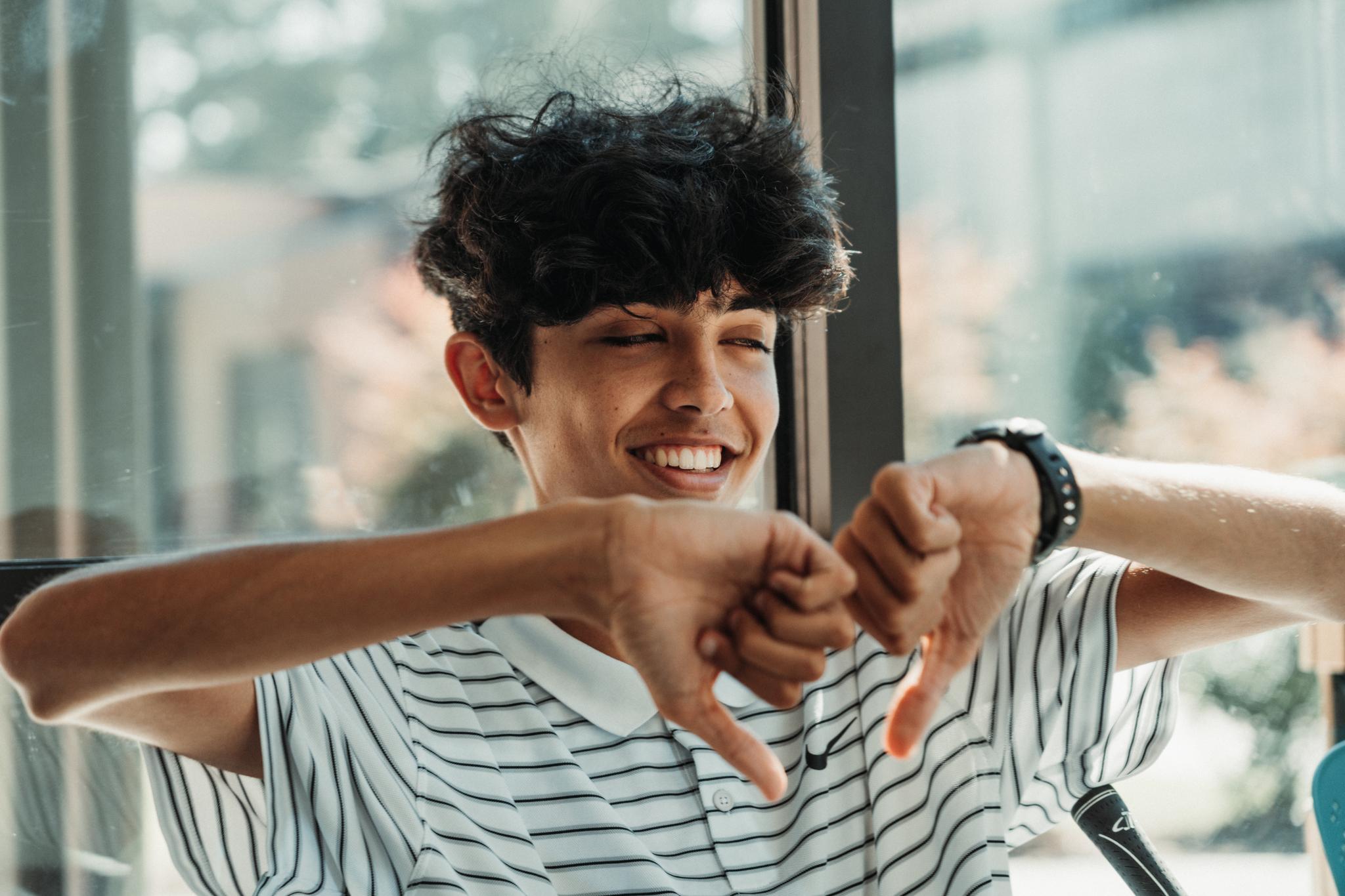 A smiling young man with curly hair is making a heart shape with his hands. He's wearing a striped shirt and a watch, and appears to be sitting in a well-lit room with windows in the background