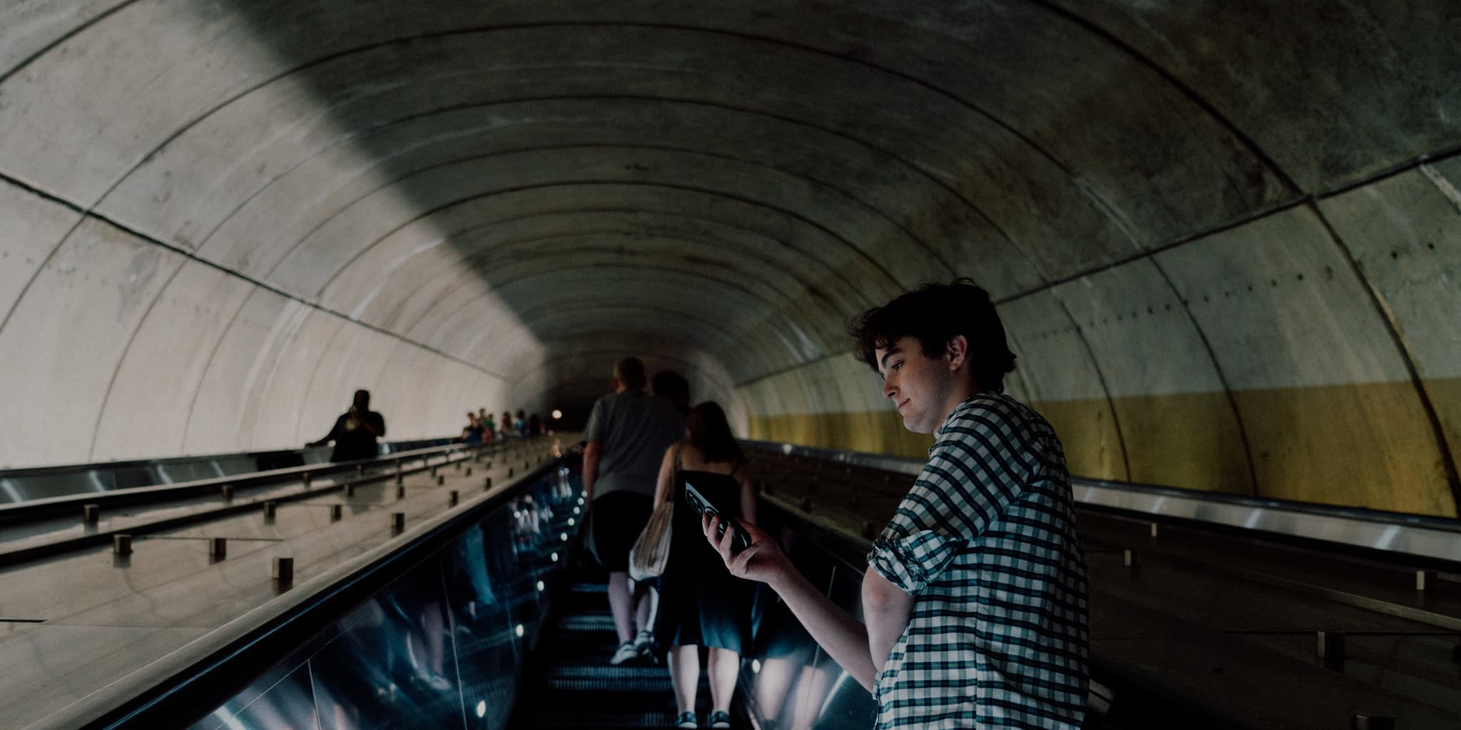 A curved metro station escalator with passengers, including a person standing on the right, descending into the tunnel